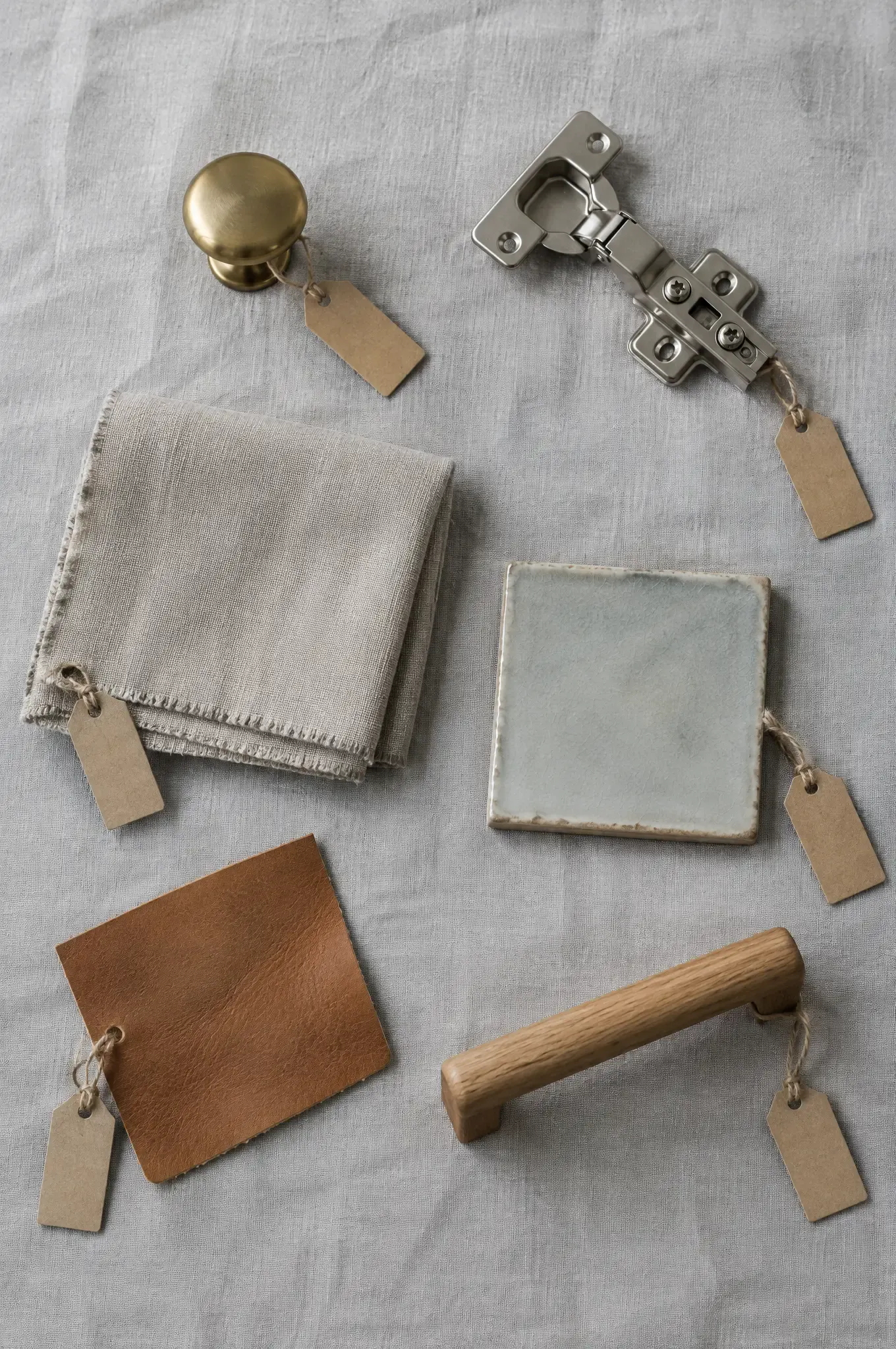 Product samples with blank tags: brass knob, steel hinge, linen, ceramic tile, leather and a wooden pull